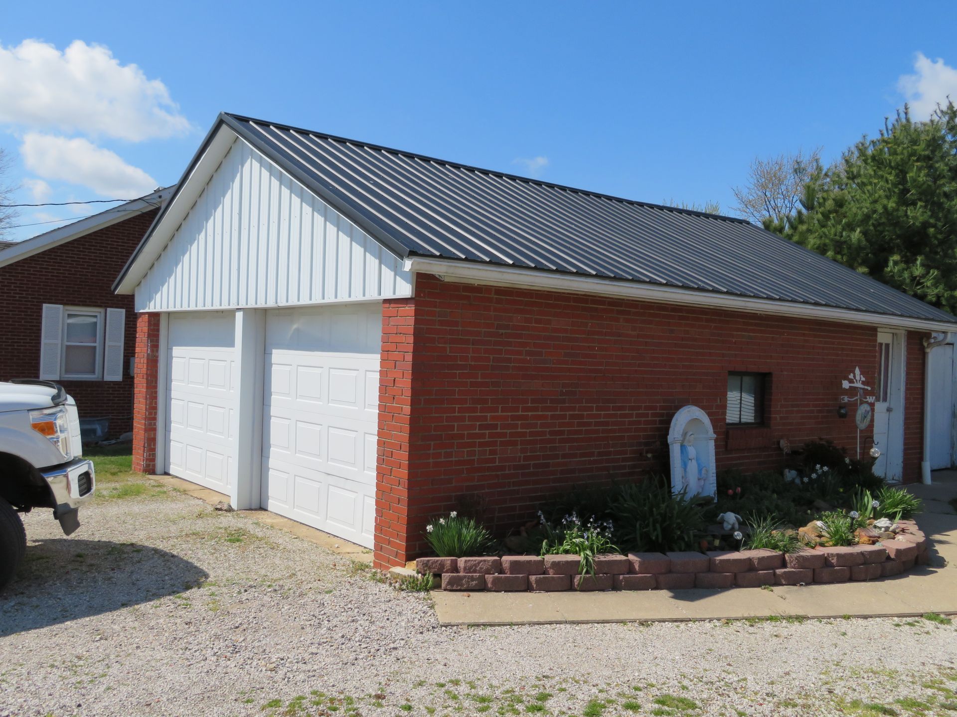 Red-brick detached garage with white doors and gray metal roof beside a gravel driveway.