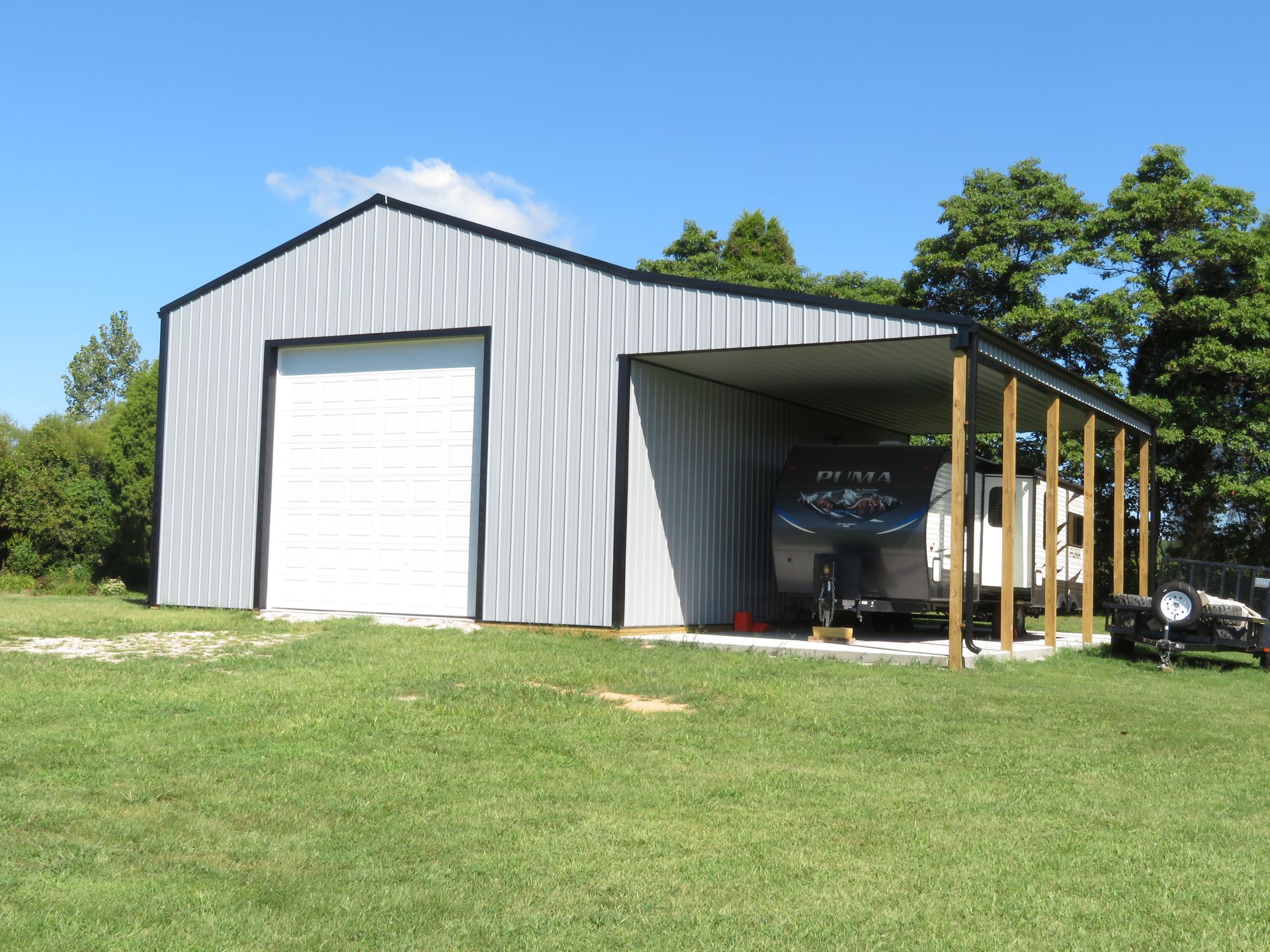 Gray metal barn with white garage door and open side bay on a grassy field under a blue sky