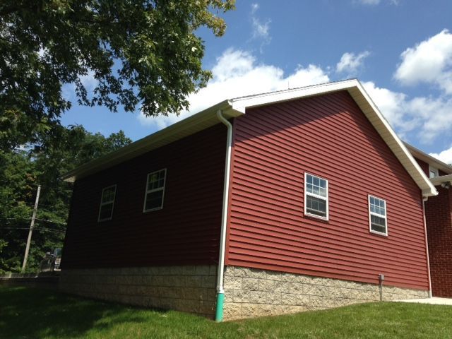 Red house with stone foundation and white-trimmed windows, viewed from the side under trees and a blue sky