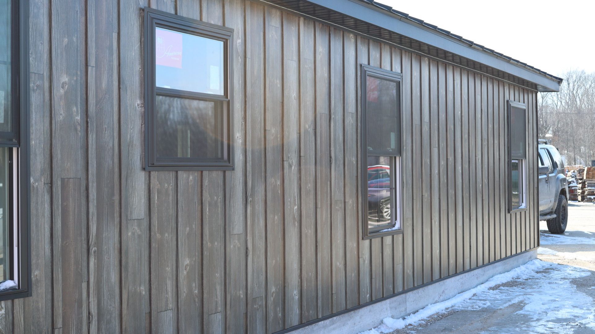 Wooden building exterior with two windows and snow along the base, viewed from the side.