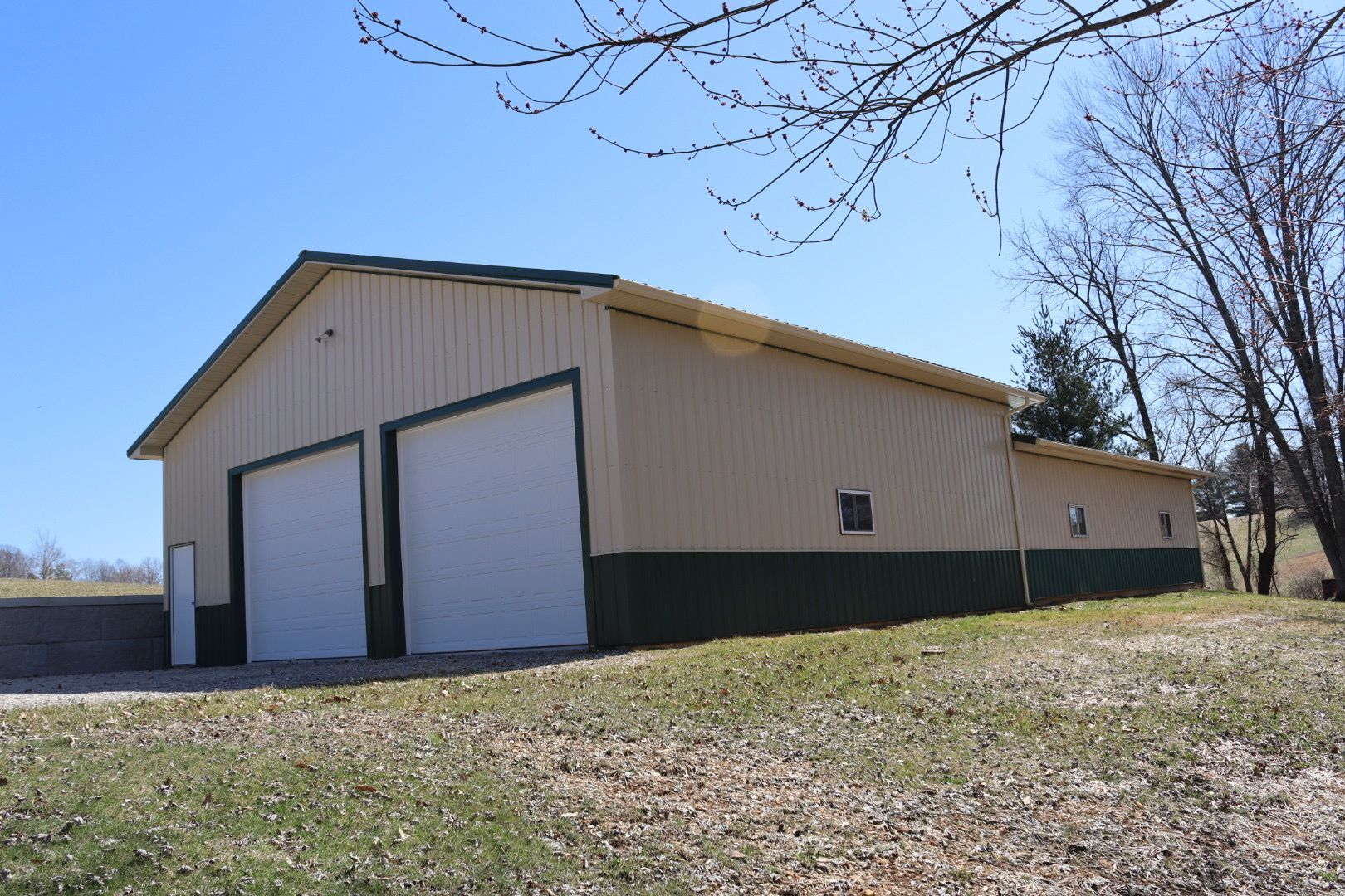 Rural beige metal barn with large white doors on a grassy lot under a clear blue sky