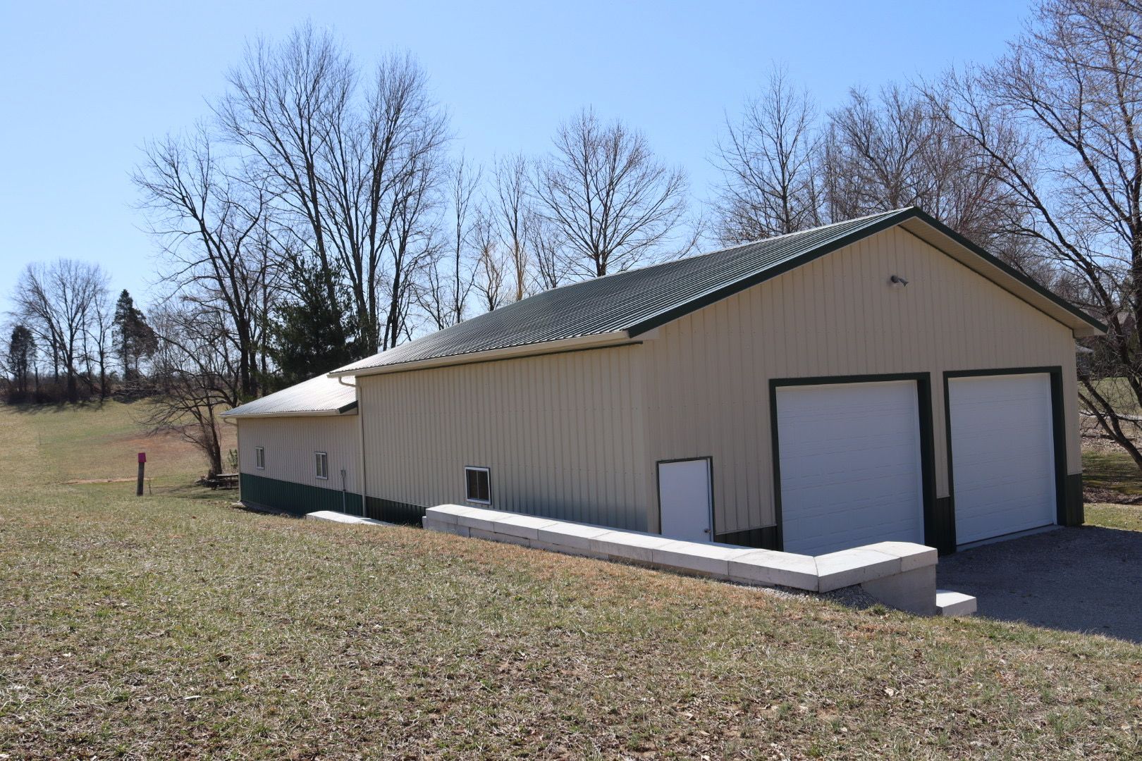 Tan metal outbuilding with two garage doors beside a grassy field and leafless trees