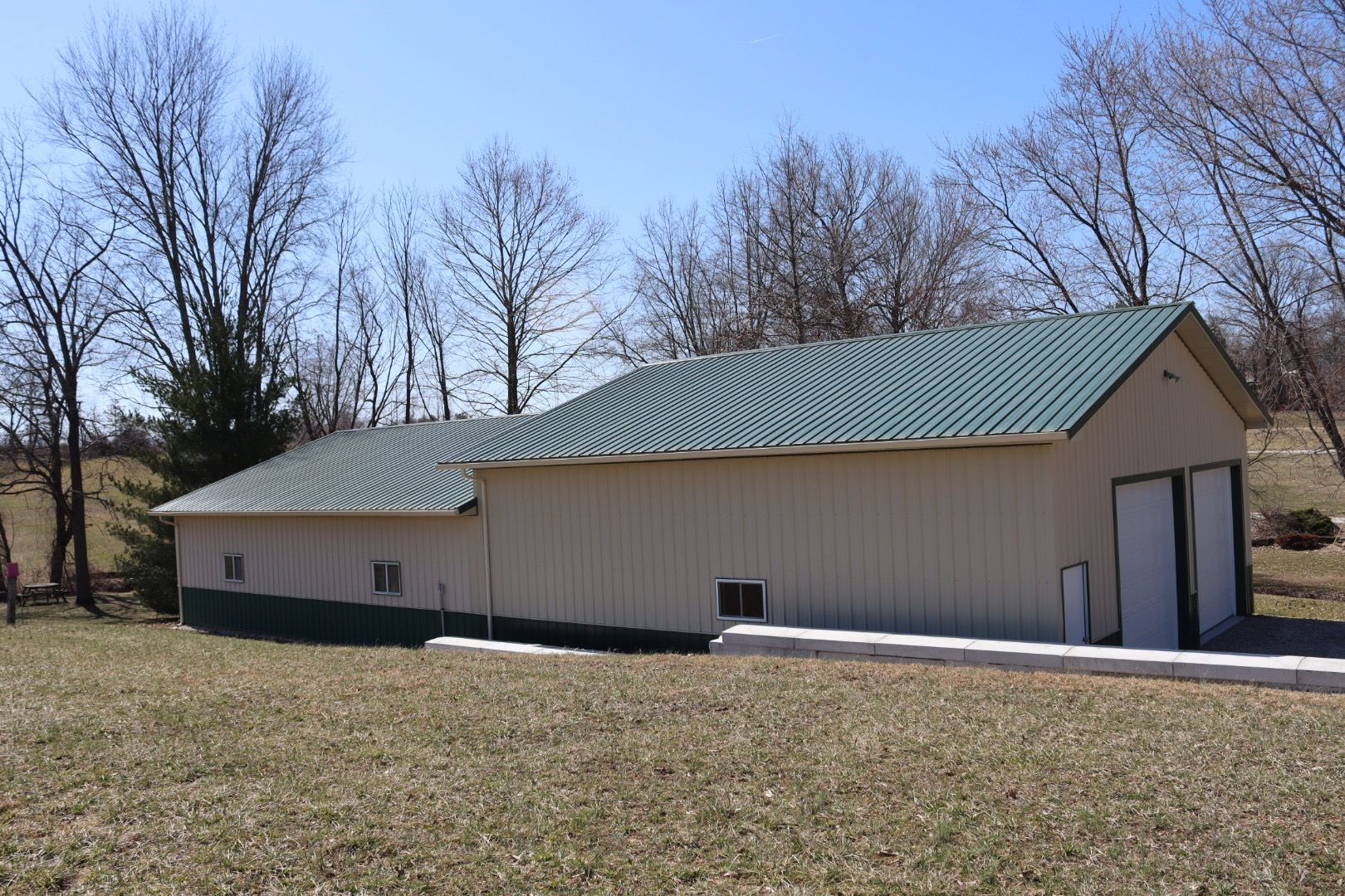 Long beige barn with green metal roof on a grassy field under a clear blue sky.
