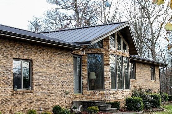 Brick house with dark metal roof and tall front windows surrounded by shrubs and trees