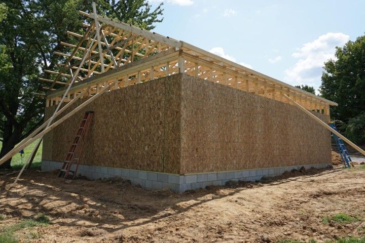 Partially built wooden house with roof trusses on a dirt lot, supported by braces under a blue sky