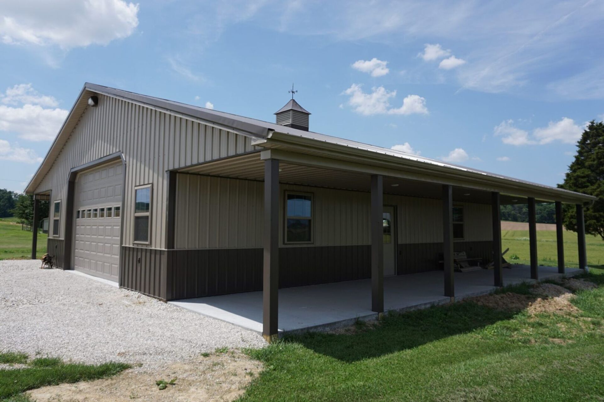 Single-story tan barn-style building with a covered porch and gravel driveway in a grassy field