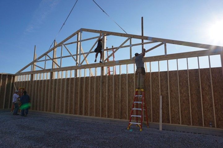 Workers framing a large wooden wall outdoors, with one person on a ladder under a bright sky.