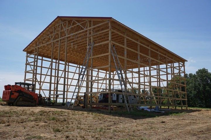 Large wooden barn frame under construction with tractors and equipment on a dirt lot, trees in background