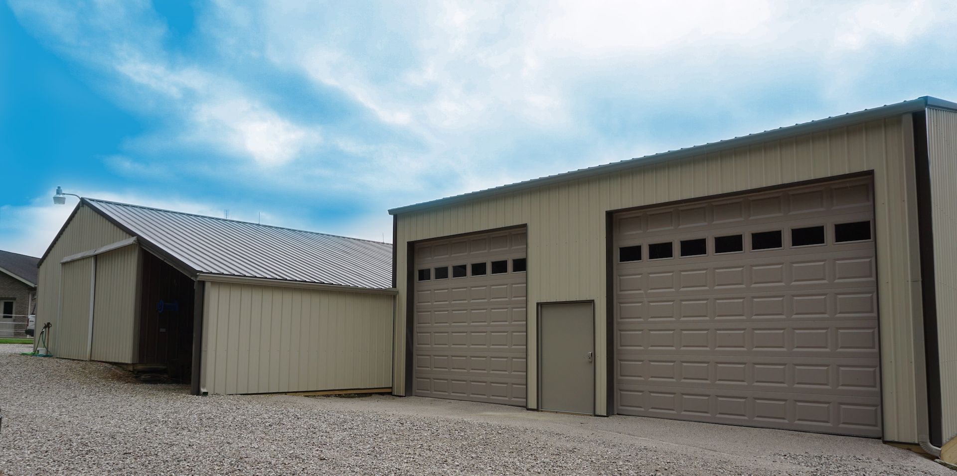 Two beige garages with metal roofs and gravel driveway under a cloudy blue sky