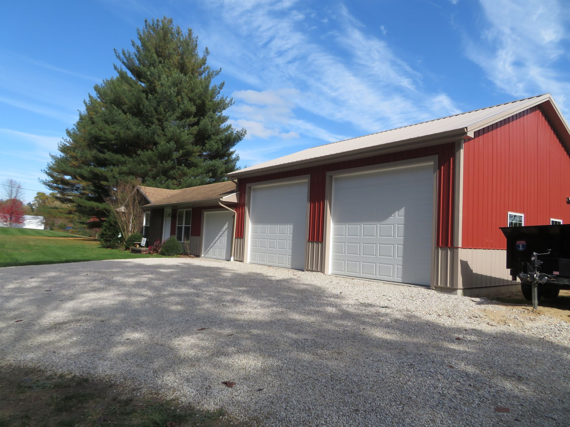 Red metal garage with three white doors beside a gravel driveway and large tree under a blue sky.