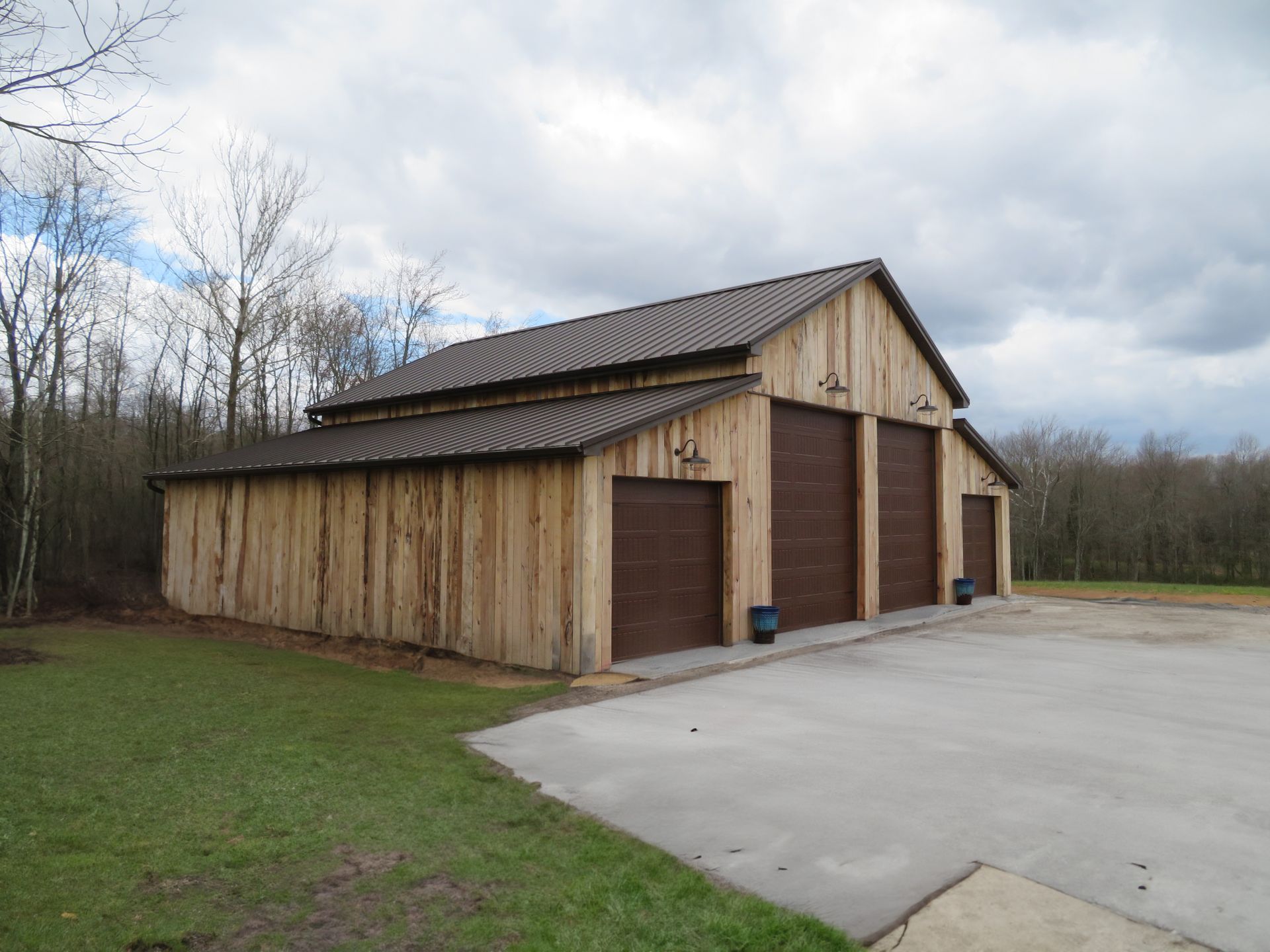Wooden barn with two large garage doors beside a concrete driveway under cloudy skies.
