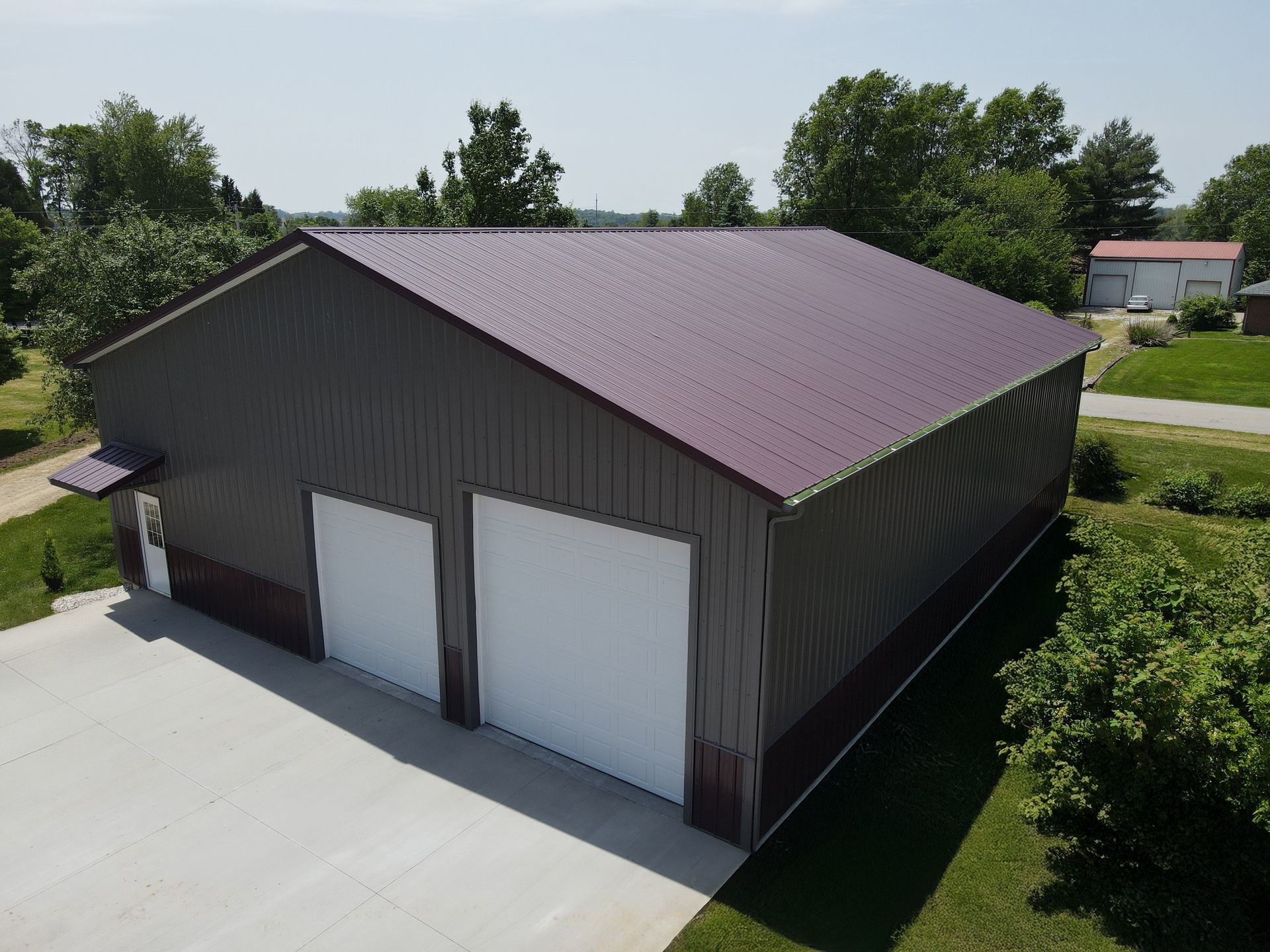 Detached gray metal garage with maroon roof and two white doors on a concrete driveway