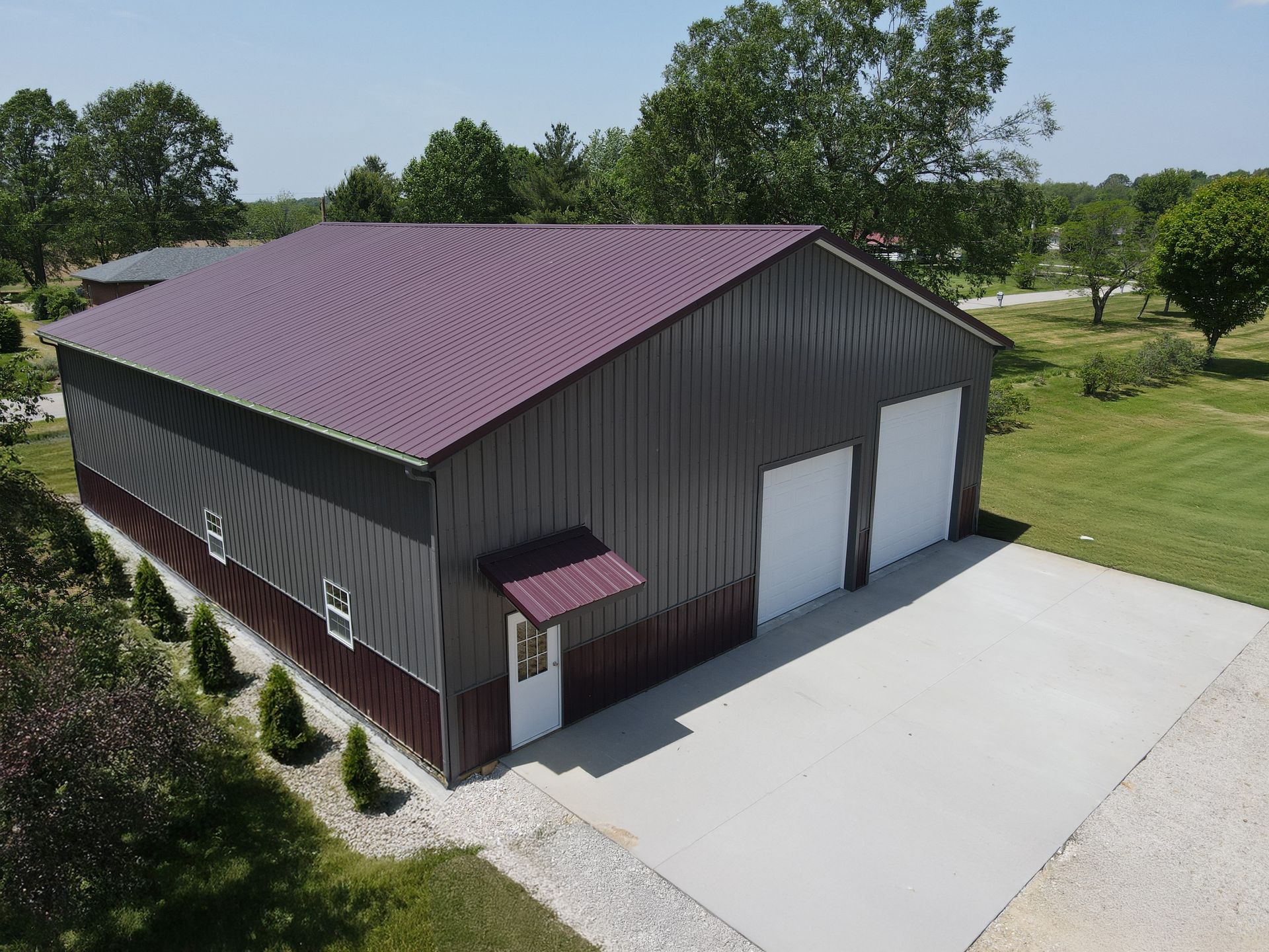 Dark metal barn with maroon roof and white garage doors beside a concrete driveway.