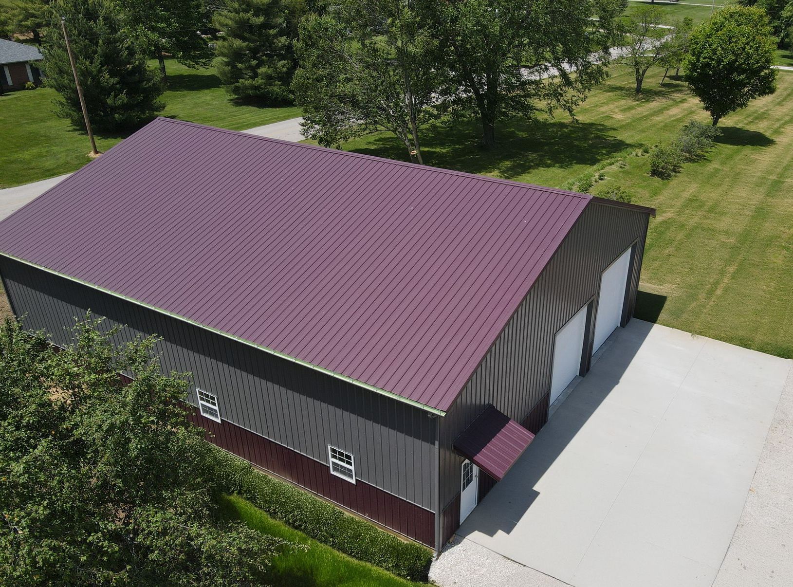 Aerial view of a large barn with a purple roof beside a paved driveway and grassy trees.