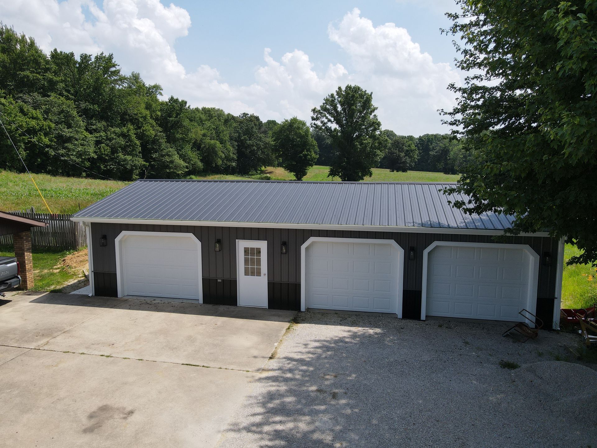 Four-car garage with dark roof and three white doors, set beside a gravel driveway and trees.