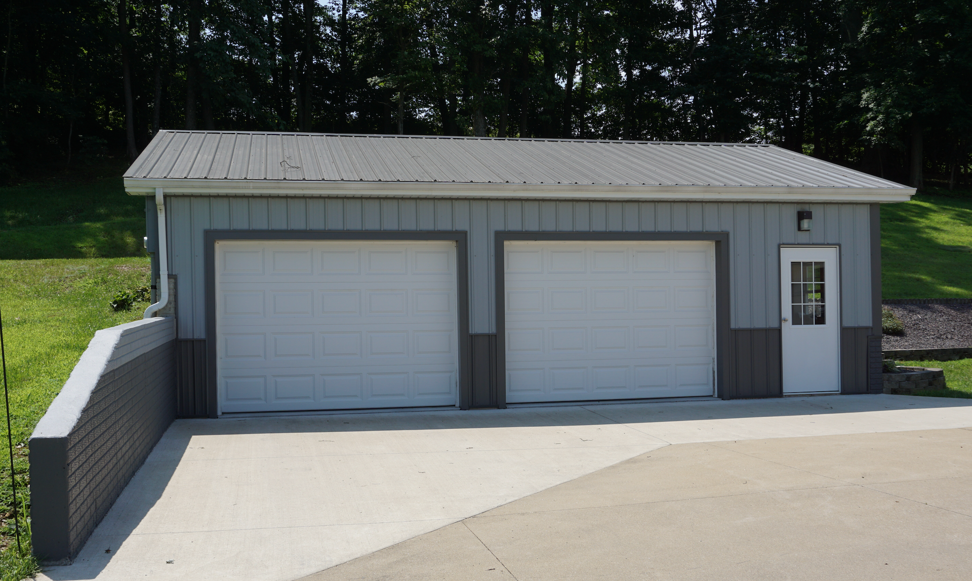 Two-car garage with gray siding, white doors, and a concrete driveway beside a wooded area