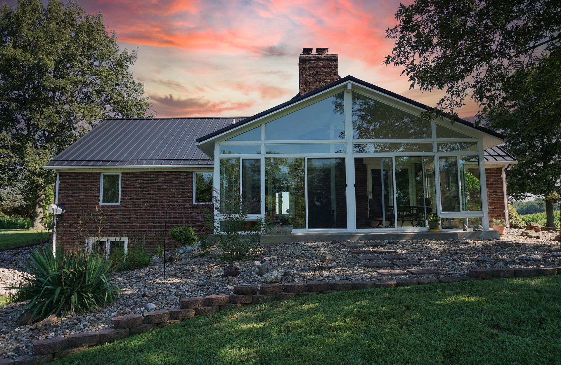 Brick house with a glass sunroom at sunset, surrounded by trees and a landscaped yard