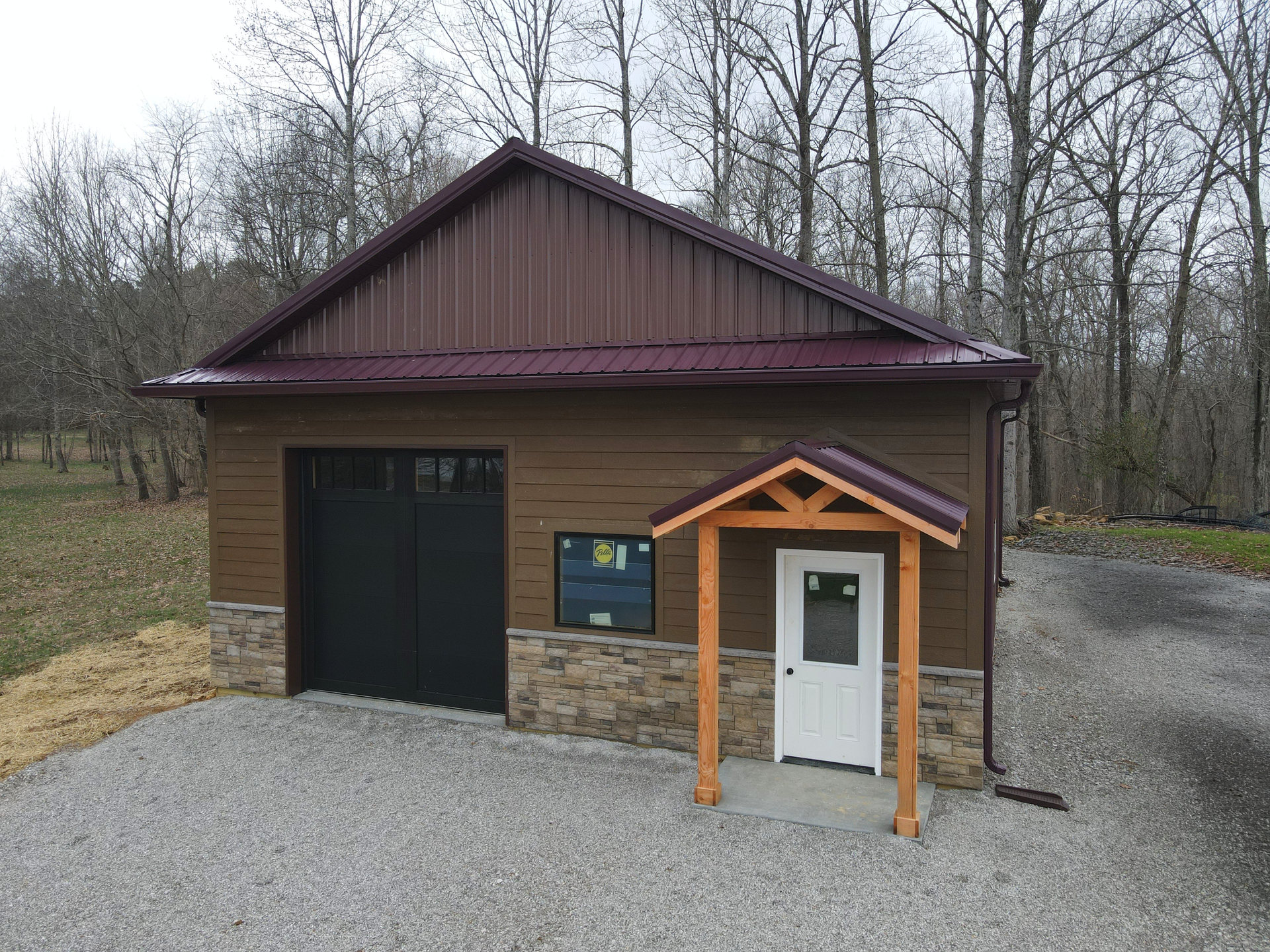 Small brown garage with stone base and black door beside a white entry door and gravel driveway in a wooded area