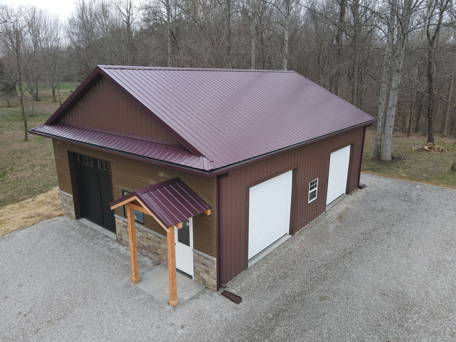 Brown outbuilding with a red metal roof, two white garage doors, and a small covered entrance in a wooded area