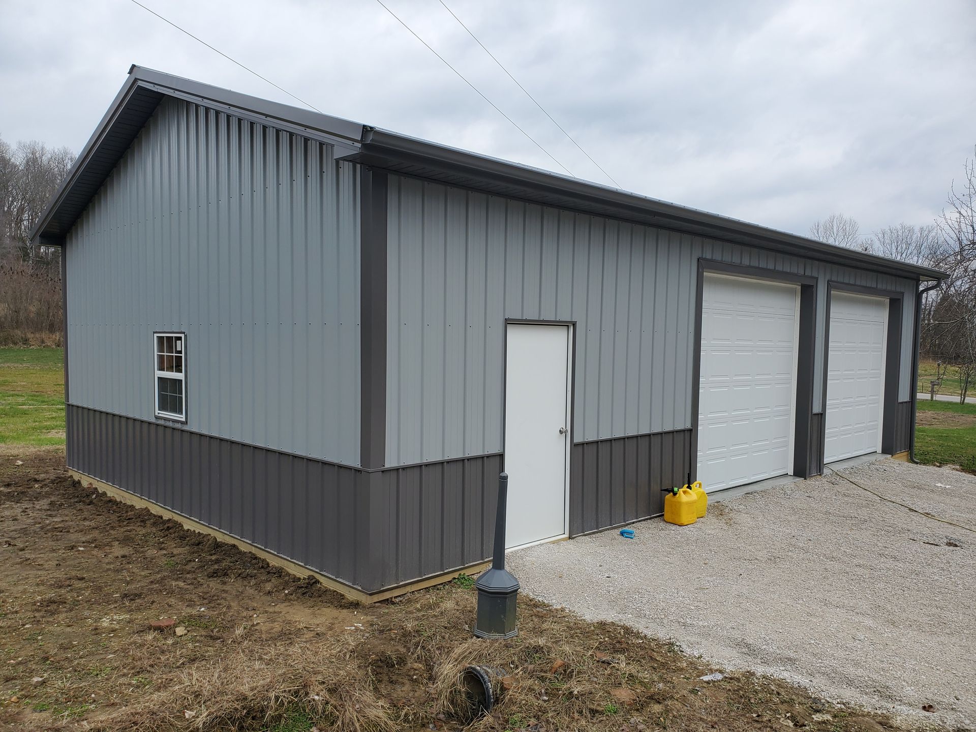Gray metal garage with white doors and gravel driveway under cloudy sky