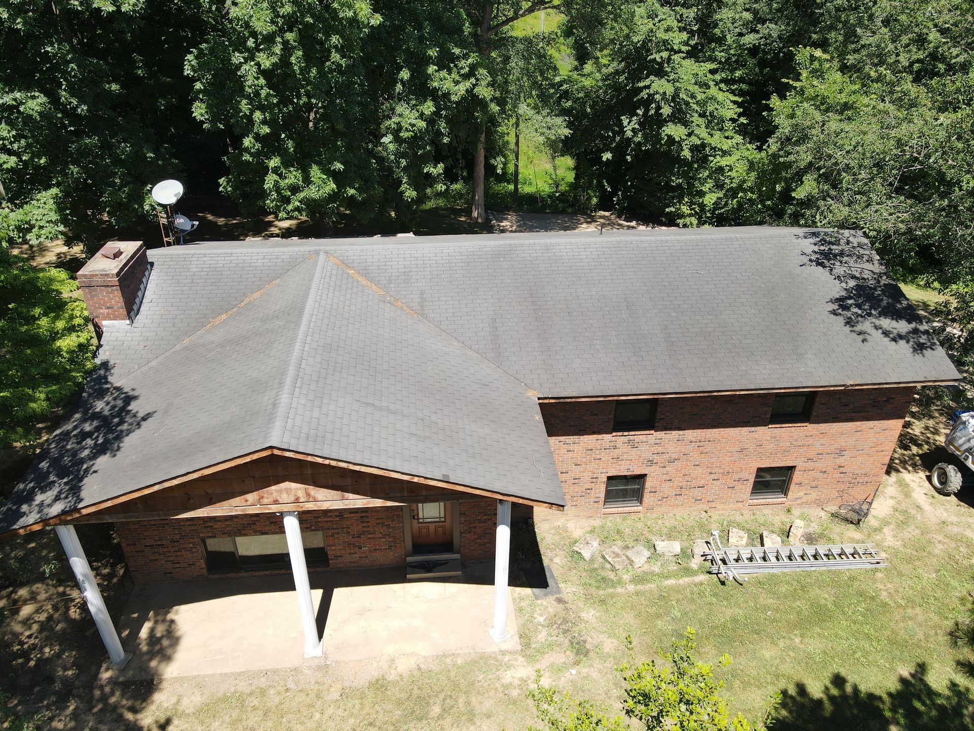 Aerial view of a brick house with a dark roof, porch, and surrounding trees and lawn