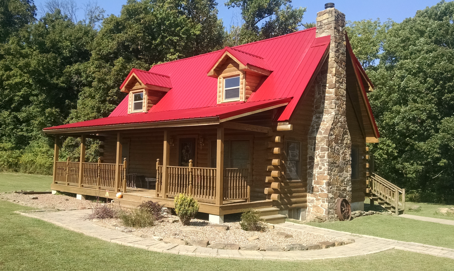Rustic log cabin with bright red roof and stone chimney in a grassy wooded setting