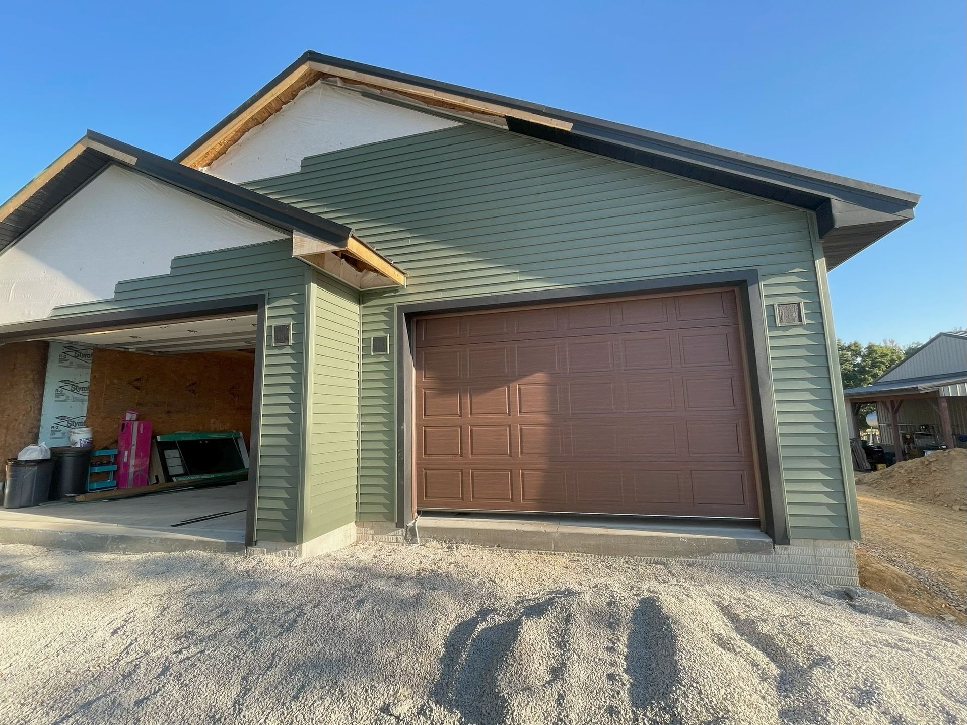 Detached house exterior with green siding, brown garage door, and gravel driveway under a blue sky