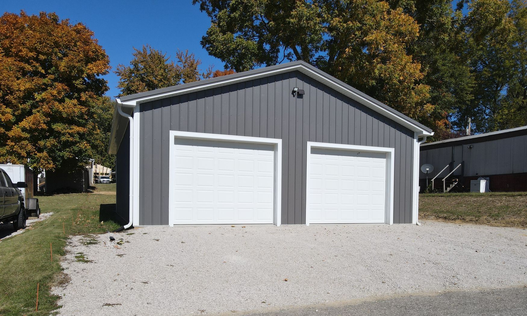 Gray metal garage with two white doors on a gravel driveway, surrounded by autumn trees