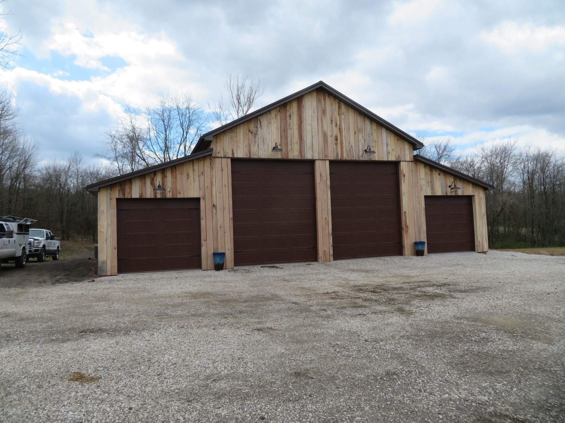 Rustic wooden barn with three brown garage doors on a gravel lot under a cloudy sky
