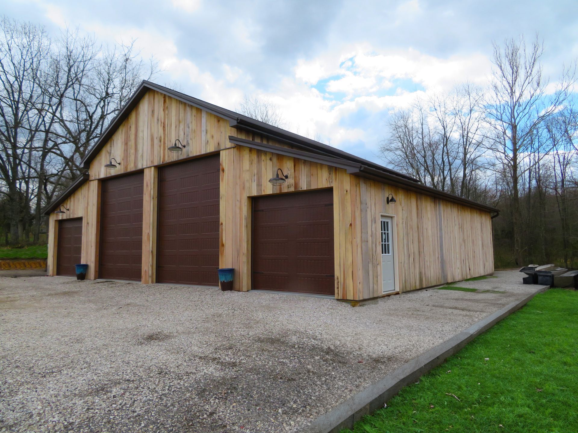 Wood-sided barn with three brown garage doors on a gravel driveway beside trees and grass