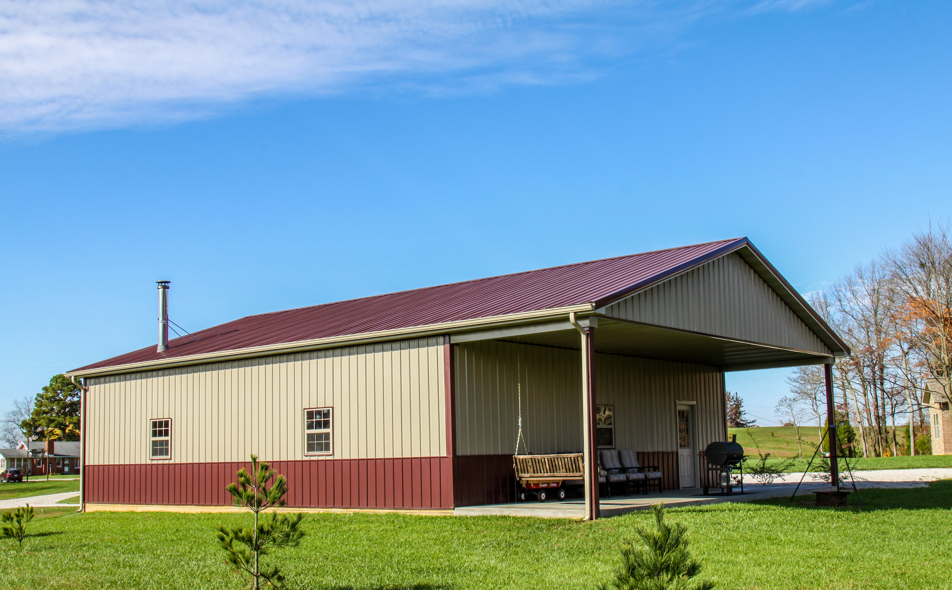 Tan and maroon metal barn-style building with an open side and grassy yard under a blue sky