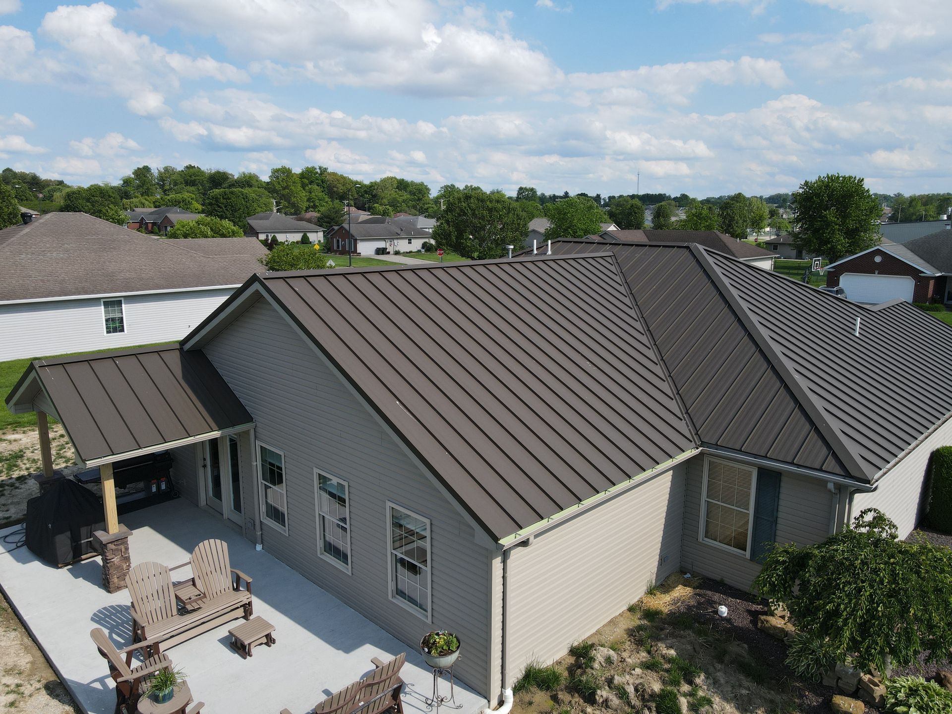 Aerial view of a gray-roofed house with a patio and backyard, surrounded by neighboring homes and trees.