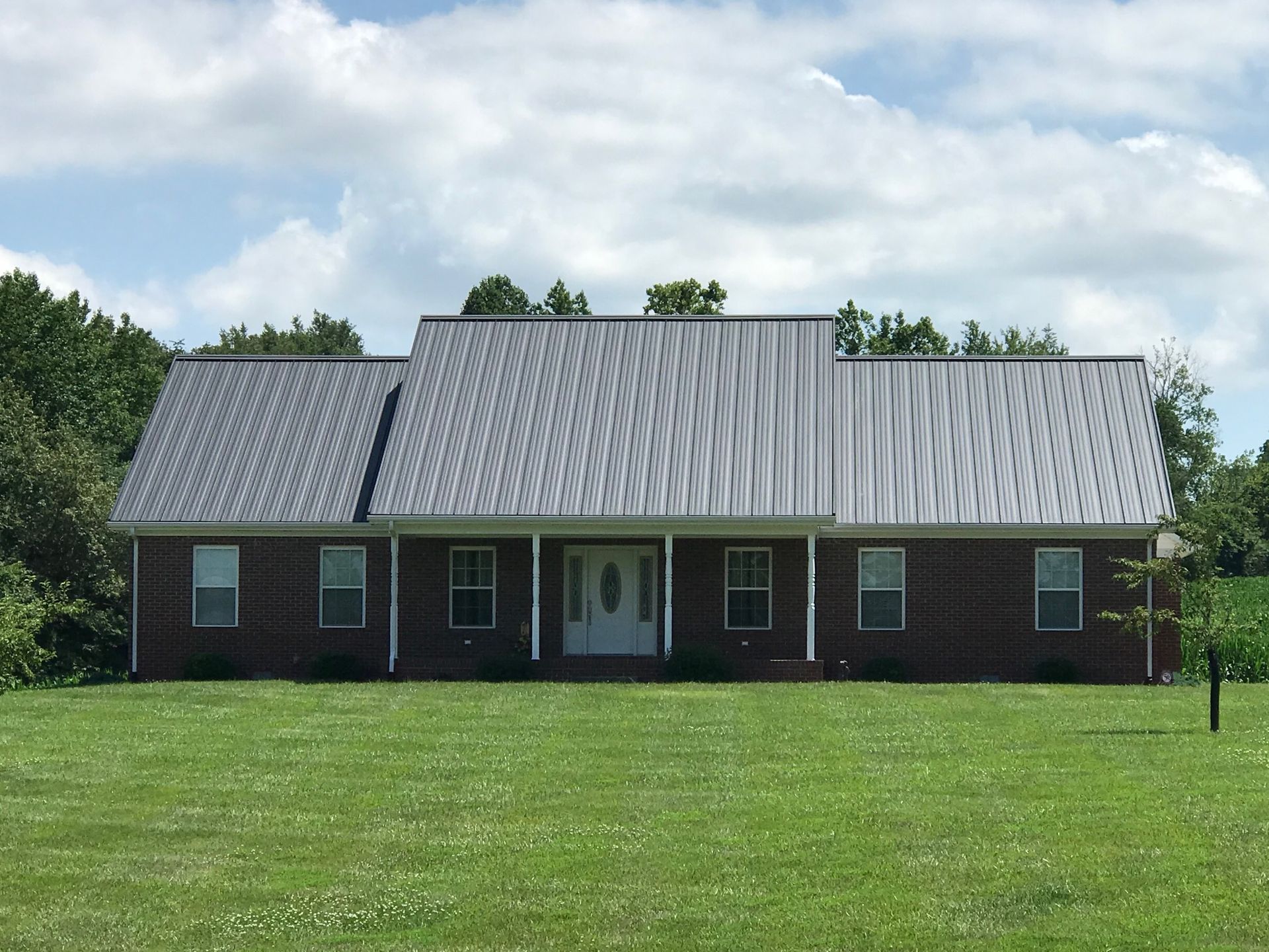 Single-story brick house with a gray metal roof and white front door, set behind a green lawn under a cloudy sky