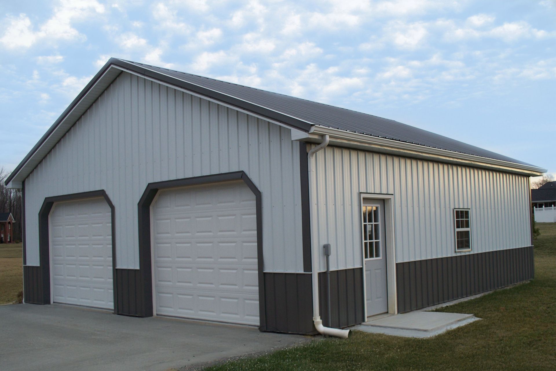 White metal garage building with two roll-up doors and a side entrance on a grassy lot under a blue sky