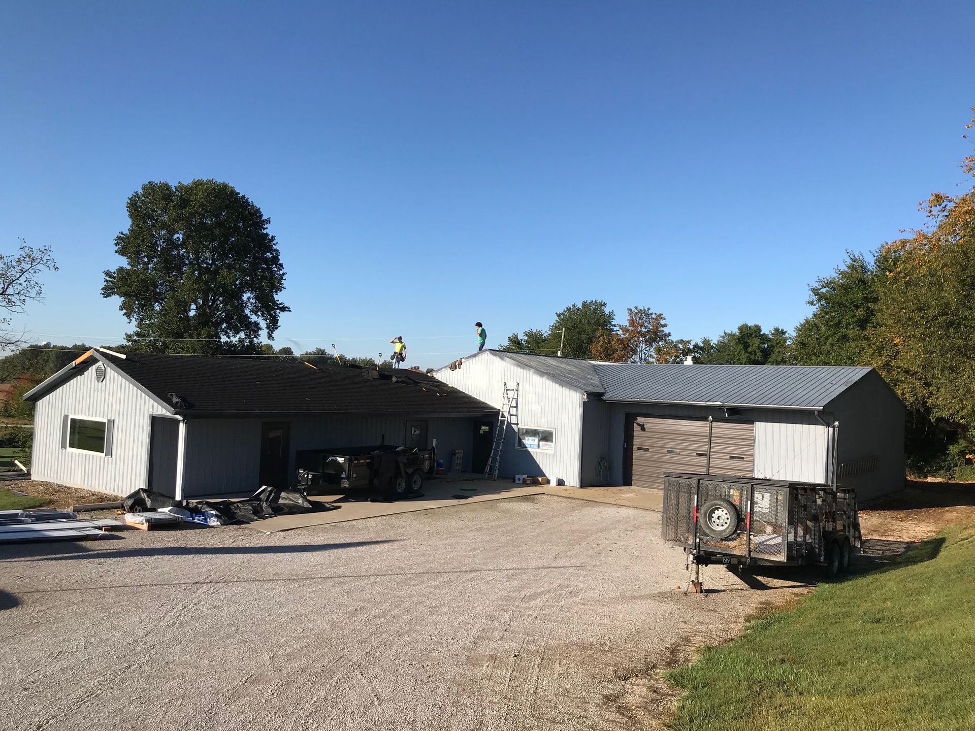Rural farmyard with white metal buildings, gravel driveway, and a utility trailer under a clear blue sky