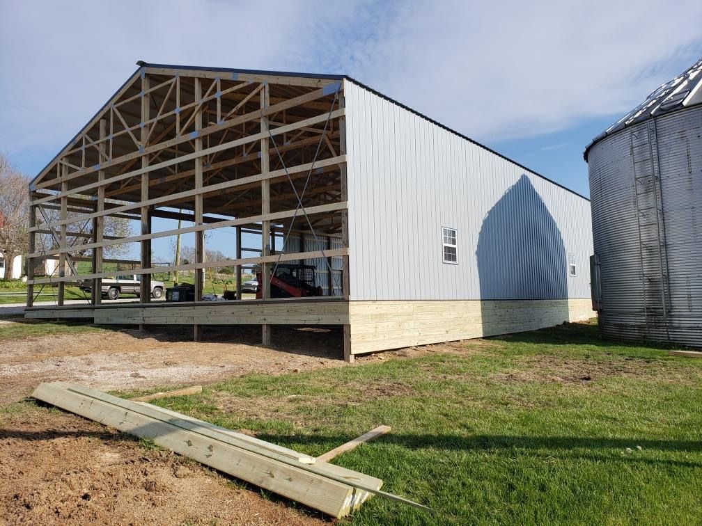 Open-sided barn under construction with white metal siding and a concrete foundation on a grassy farm.