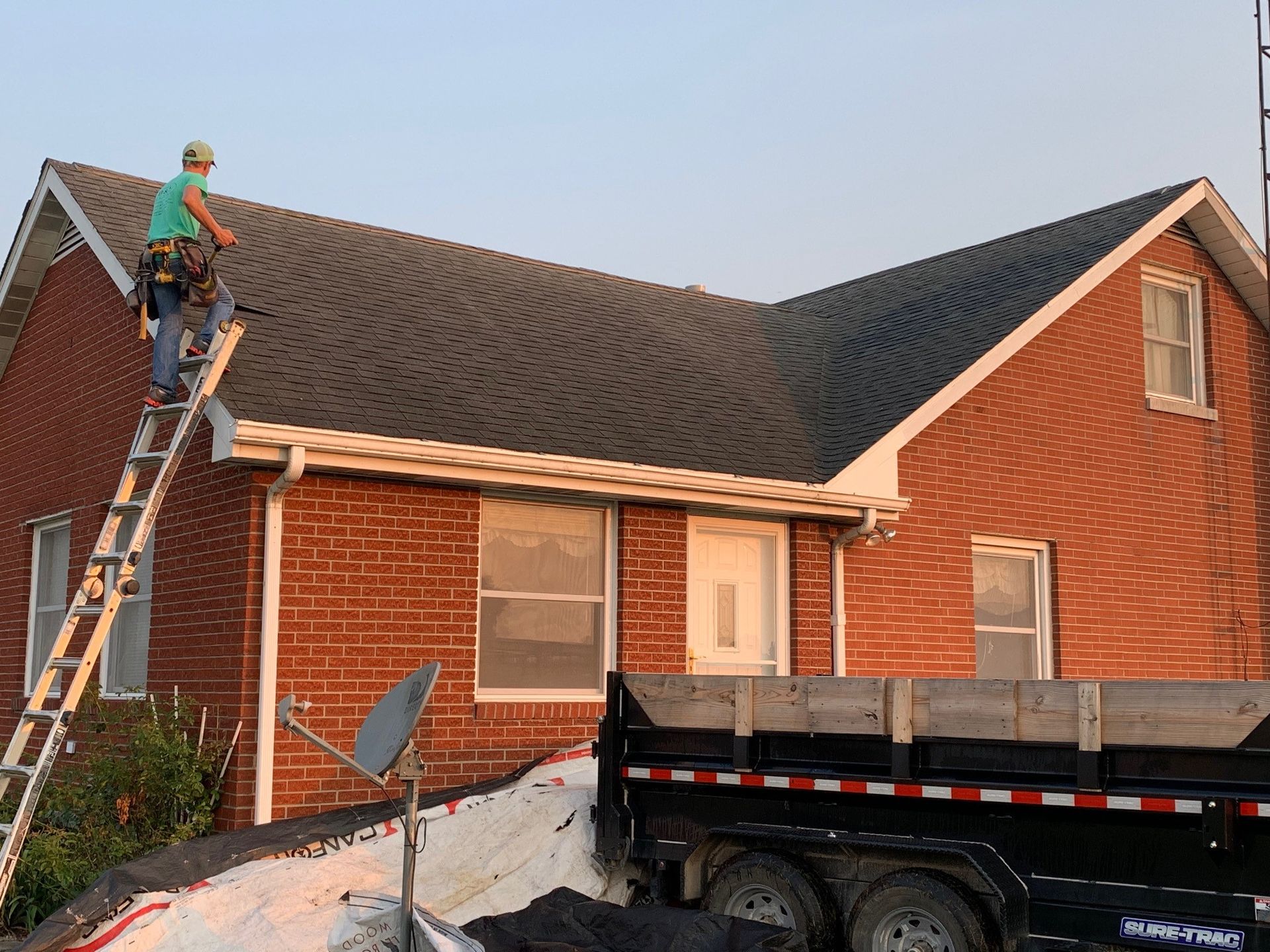 Worker on a ladder repairing a brick house roof beside a dump truck and snow-covered yard