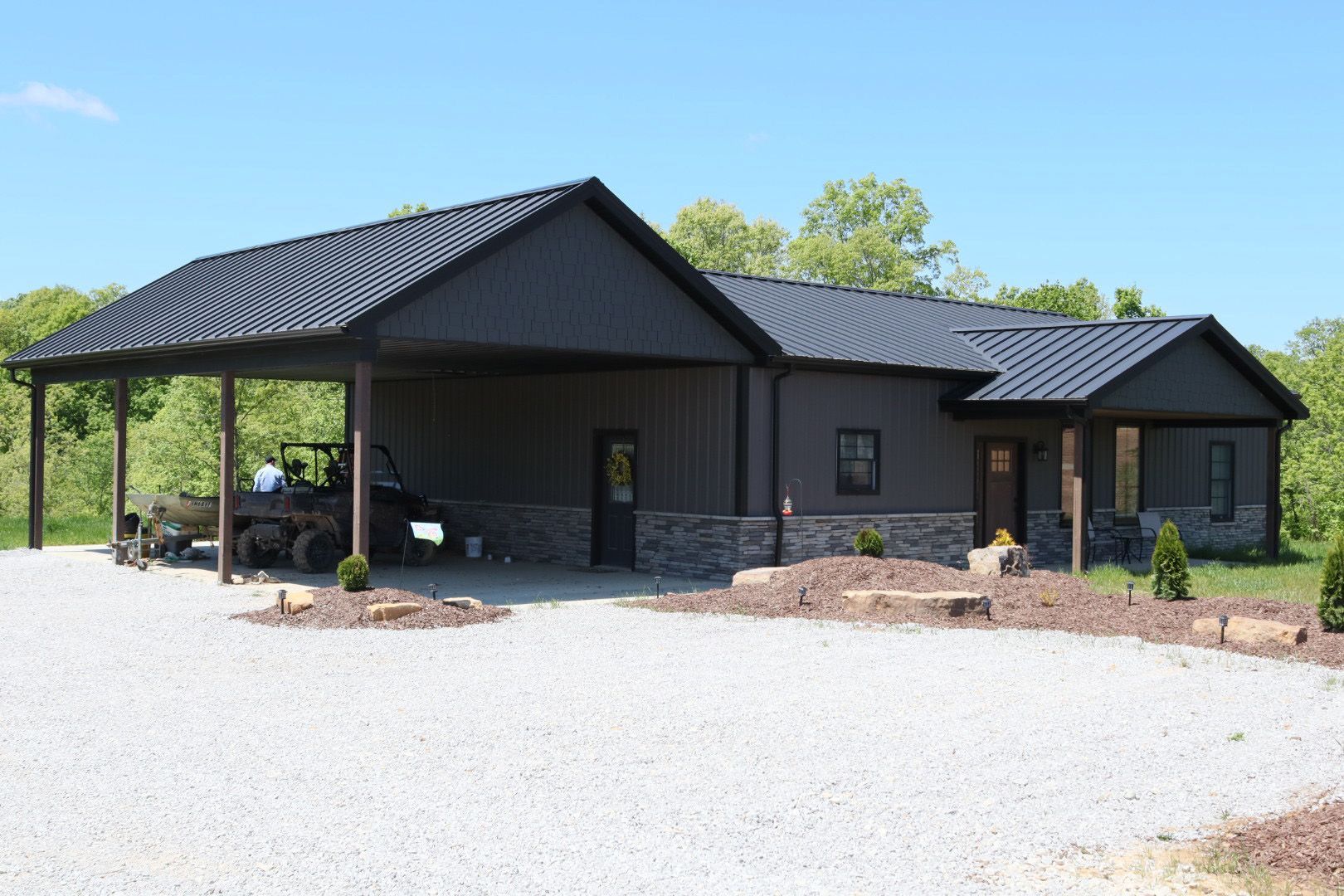 Dark gray ranch-style building with covered porch and gravel driveway, surrounded by trees and grass