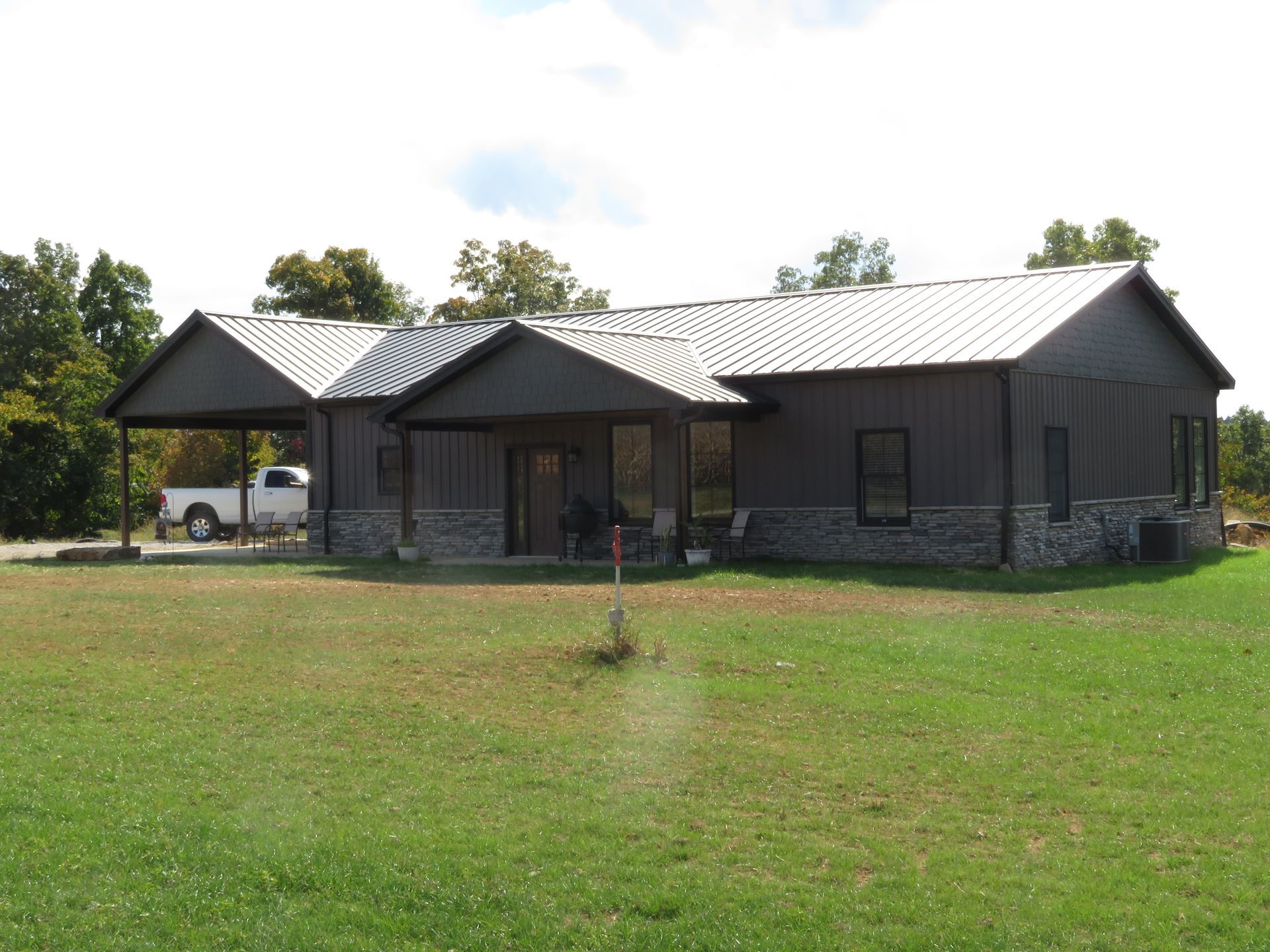 Single-story dark house with a metal roof, carport, and a large green lawn in front.