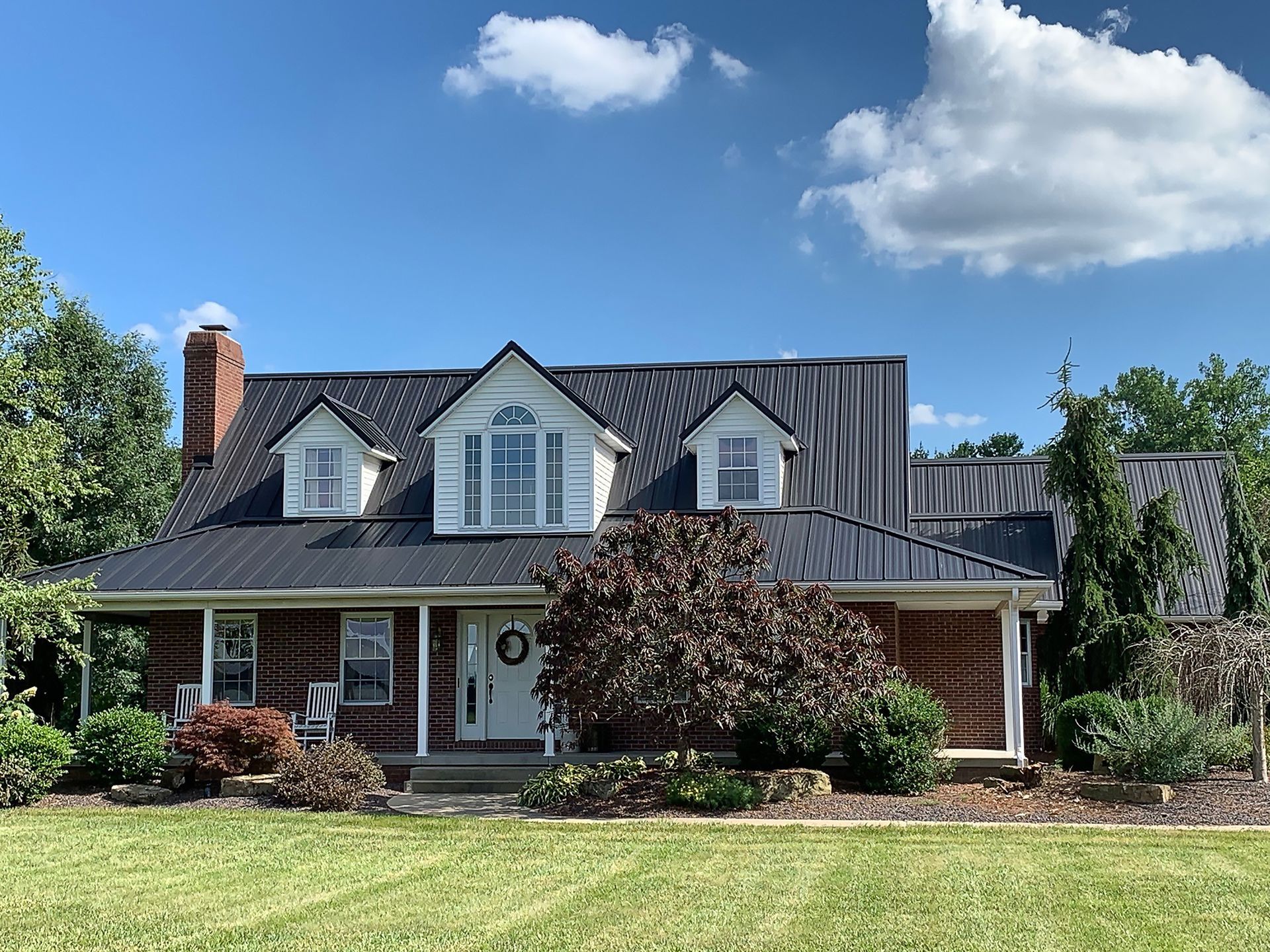 Suburban brick house with dormer windows, front porch, and landscaped yard under a blue sky