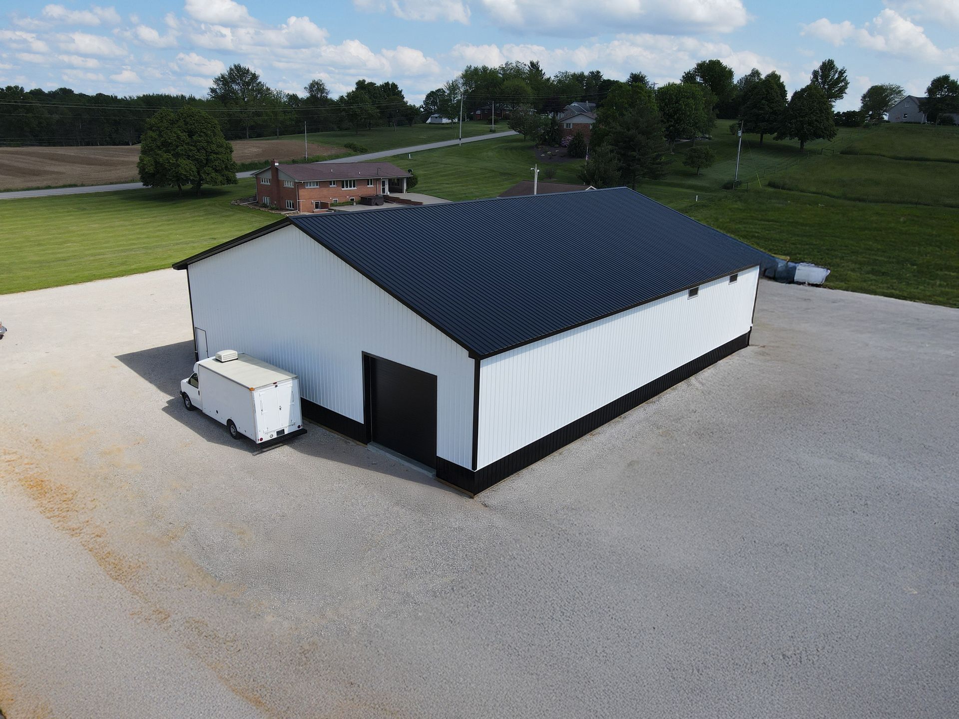 White metal warehouse with black roof in a gravel lot, viewed from above with green fields behind.