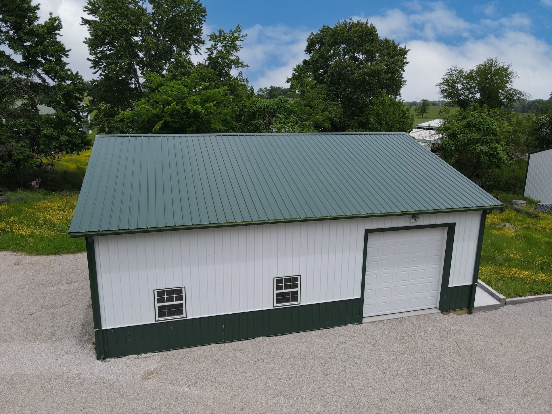 Small white shed with green roof and trim beside a gravel driveway in a grassy yard