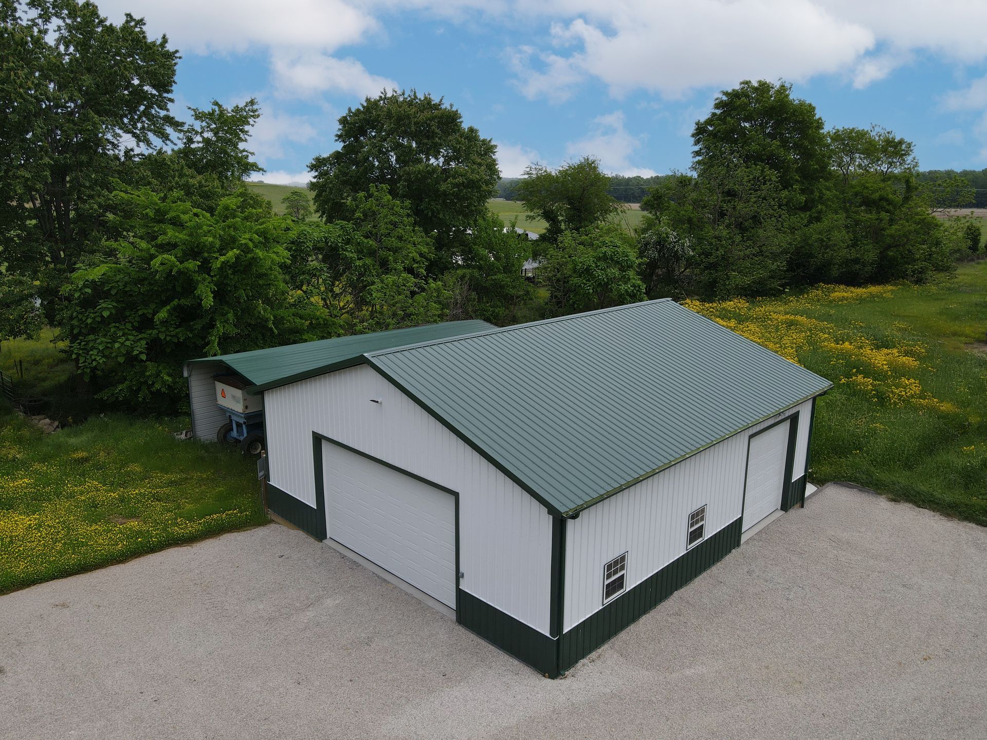 White garage with green roof on a gravel lot beside trees and wildflowers under a cloudy sky