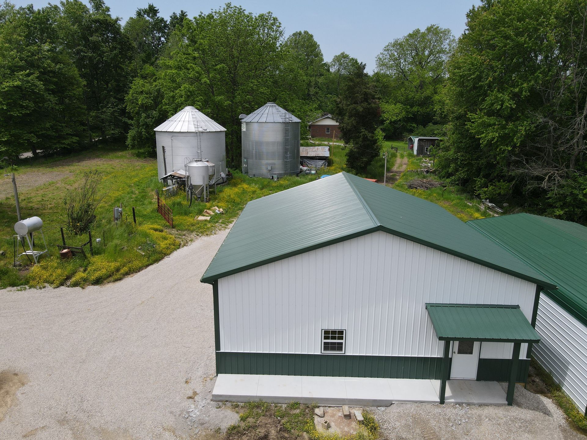 Farm buildings with green roofs beside silver grain bins and a gravel drive, surrounded by trees.