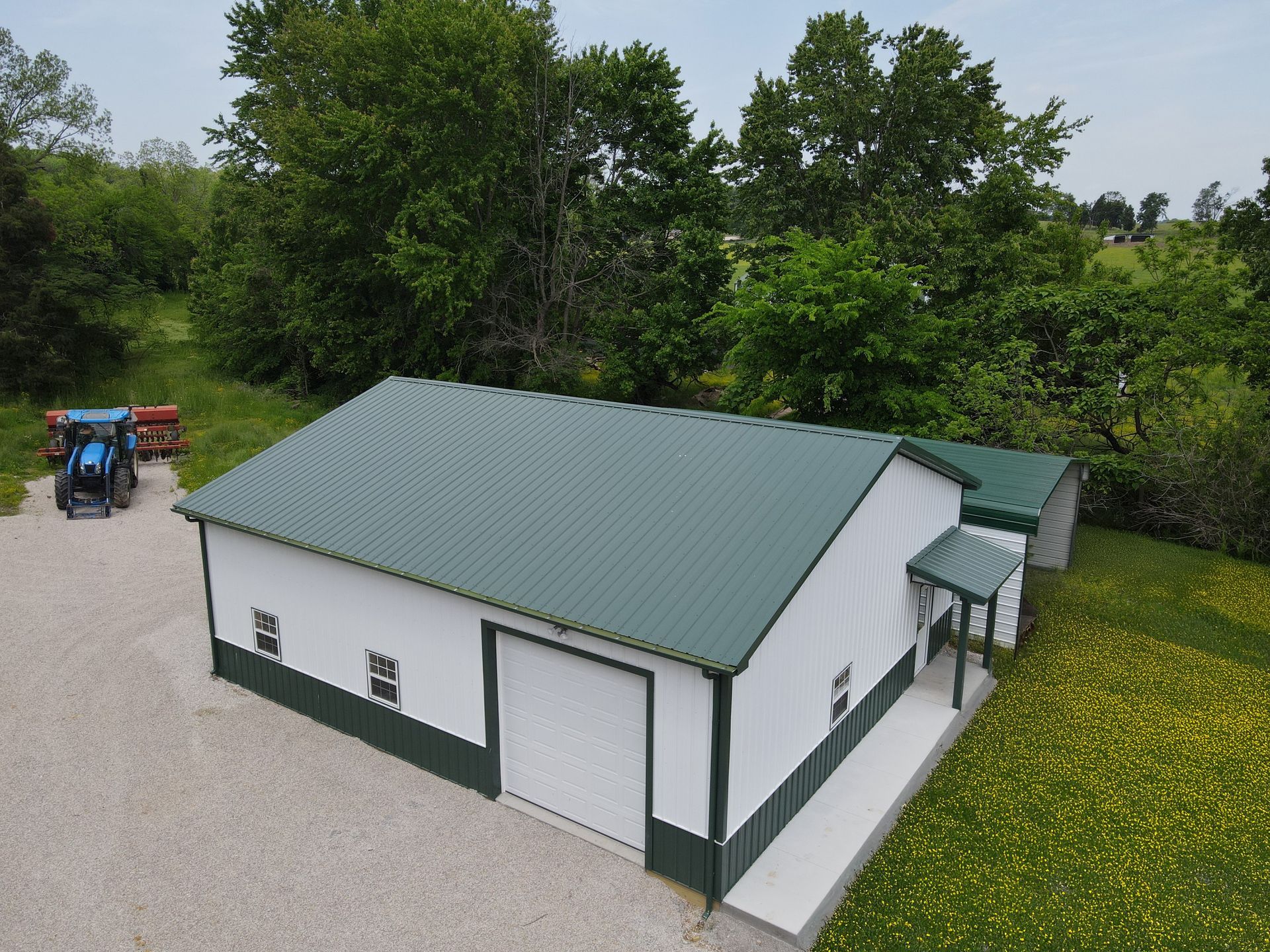 White farmhouse with green roof beside a gravel yard and trees, with a tractor in the background