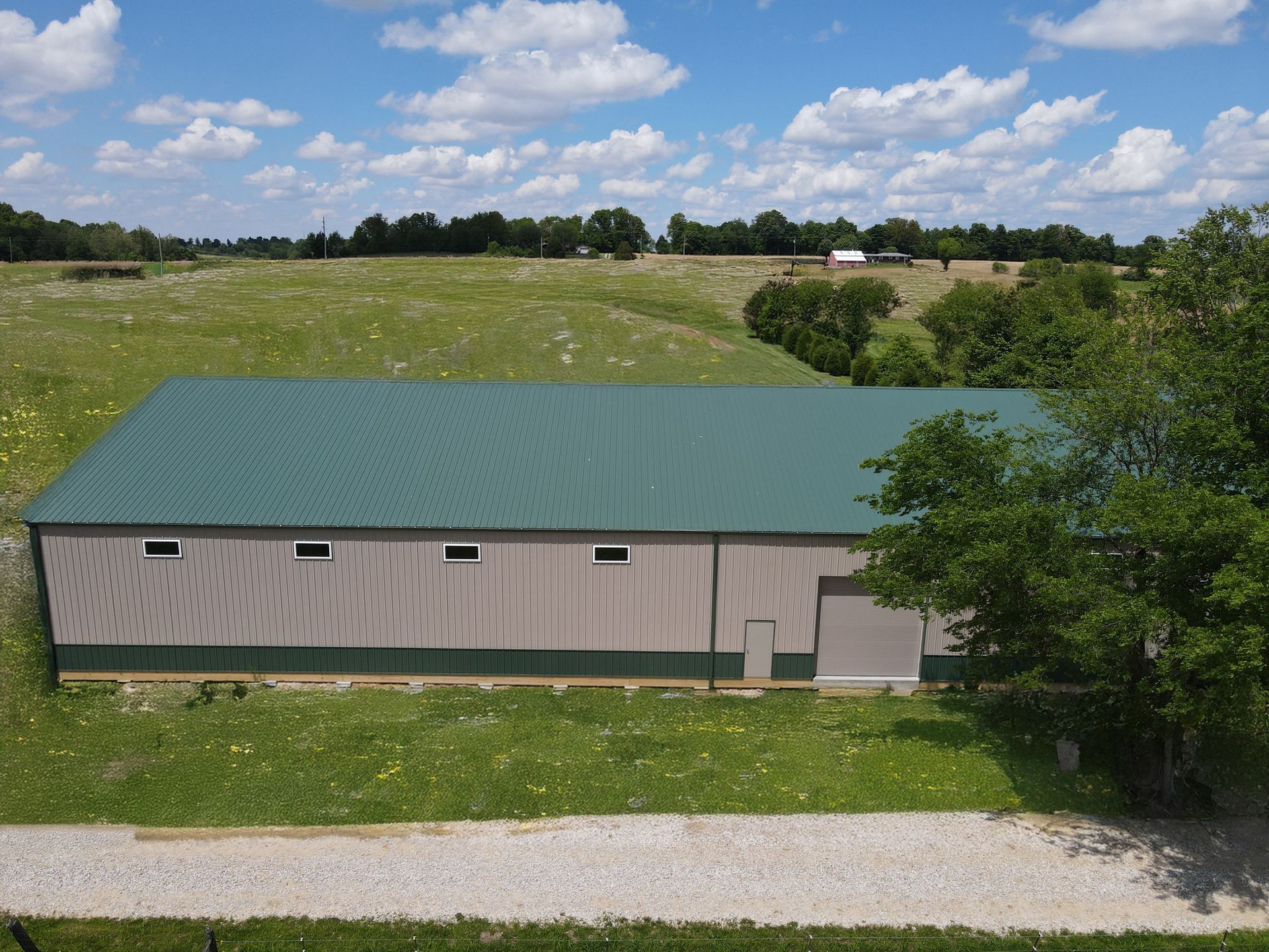 Long green-roofed barn beside a gravel road, set in a grassy field under a blue sky with clouds