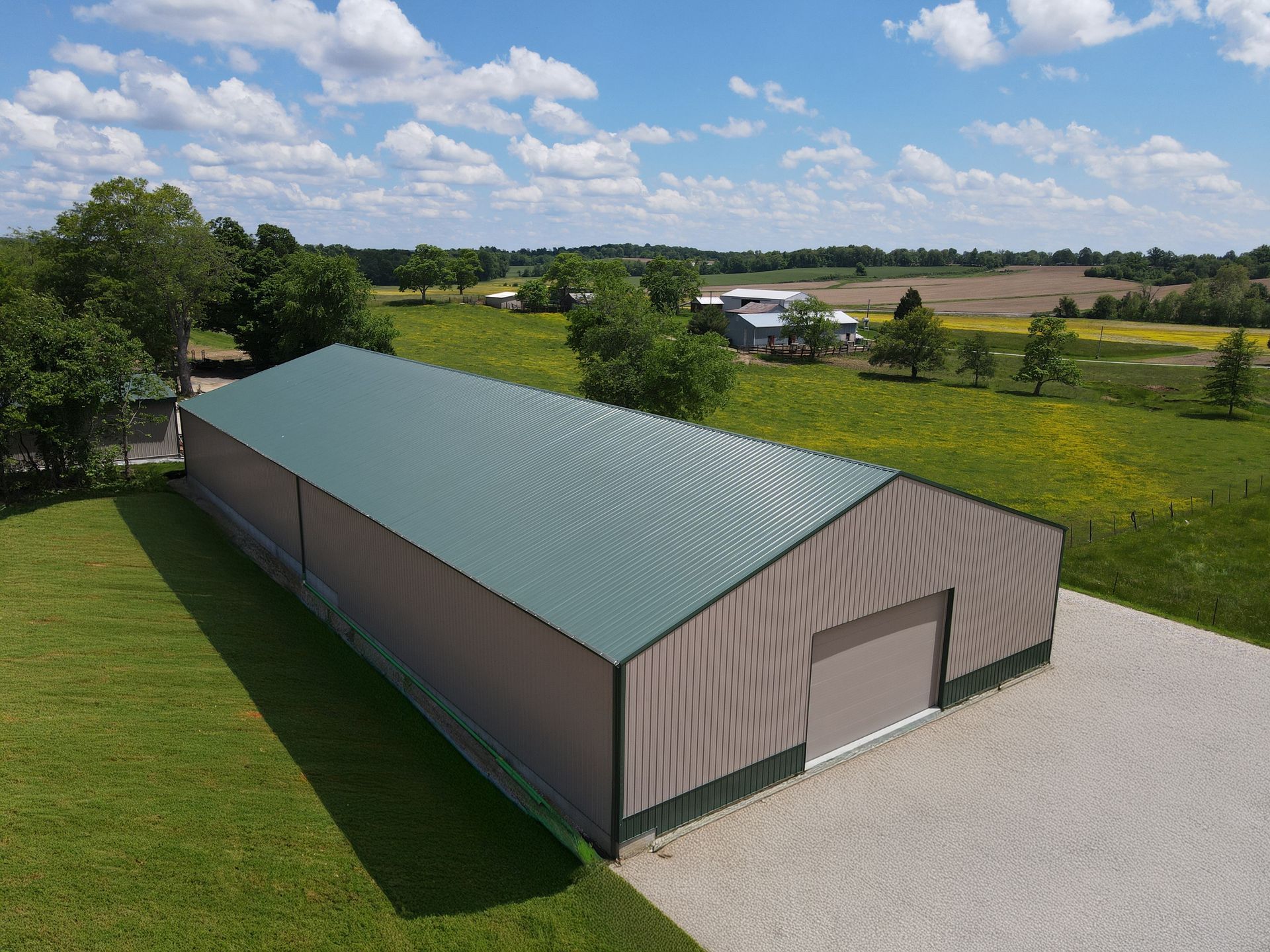Large metal barn with a green roof on a gravel lot beside grassy fields and trees under a blue sky