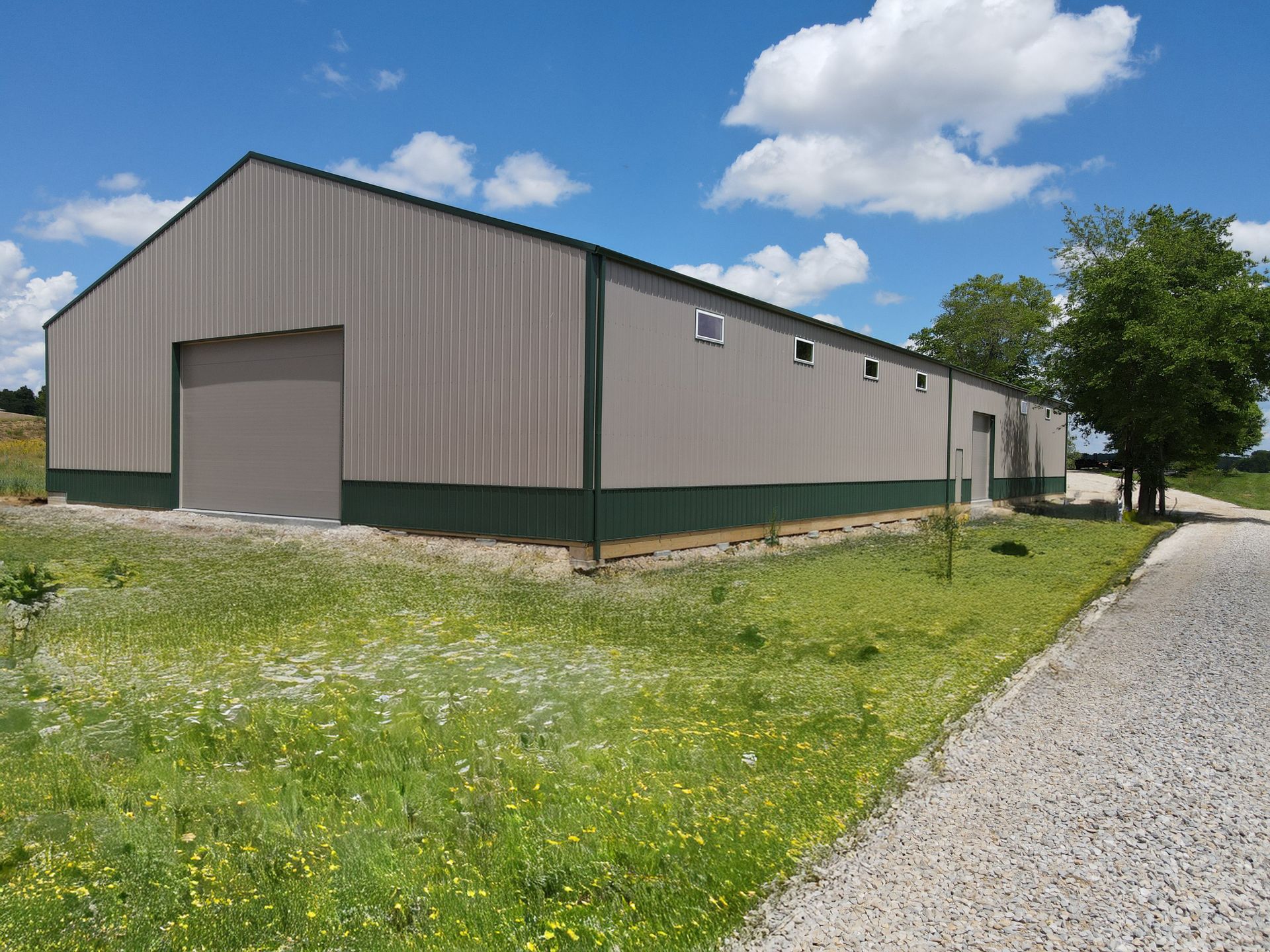 Large beige metal warehouse with green trim beside a gravel road under a blue sky