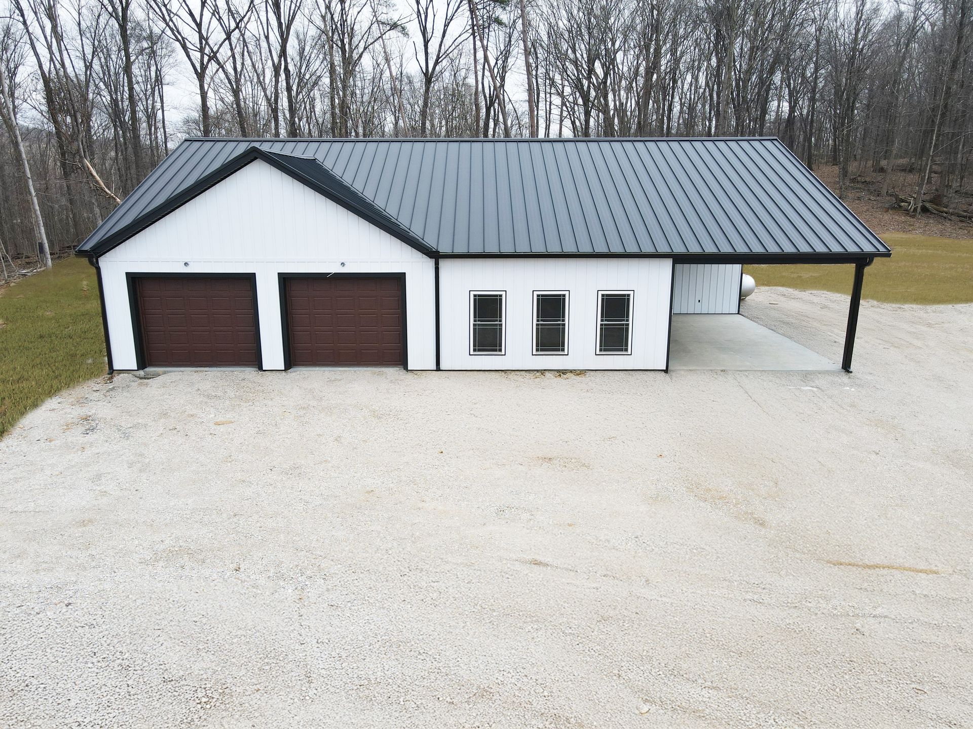 White garage with two brown doors, three windows, and a metal roof on a gravel lot by trees