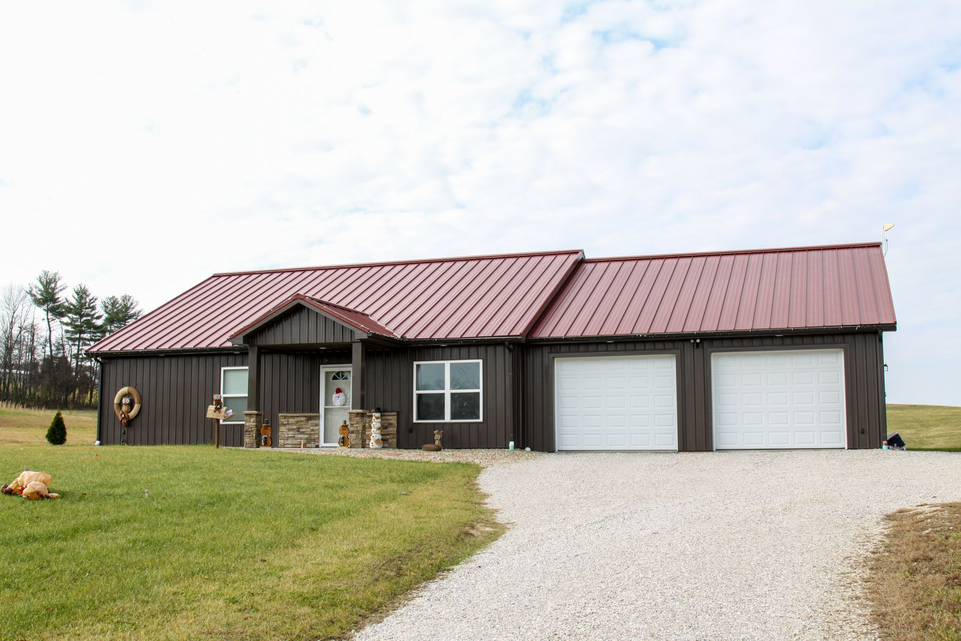 Single-story tan house with red metal roof, two garages, and gravel driveway in a grassy rural setting